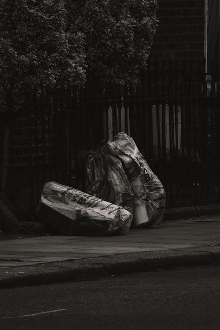 Two large black plastic rubbish bags, filled and tied, rest on the edge of a paved sidewalk near a dark wooden fence with vertical slats in an outdoor urban setting. The bags appear to contain mixed w